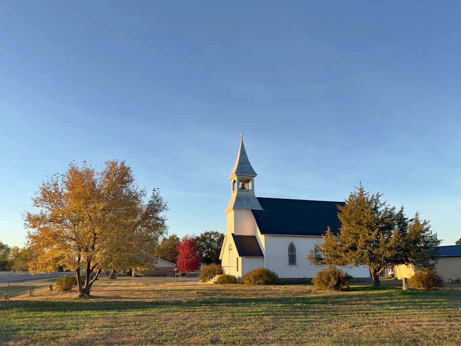 A white wood-frame church with a steeple and a dark roof, a golden maple on the lawn