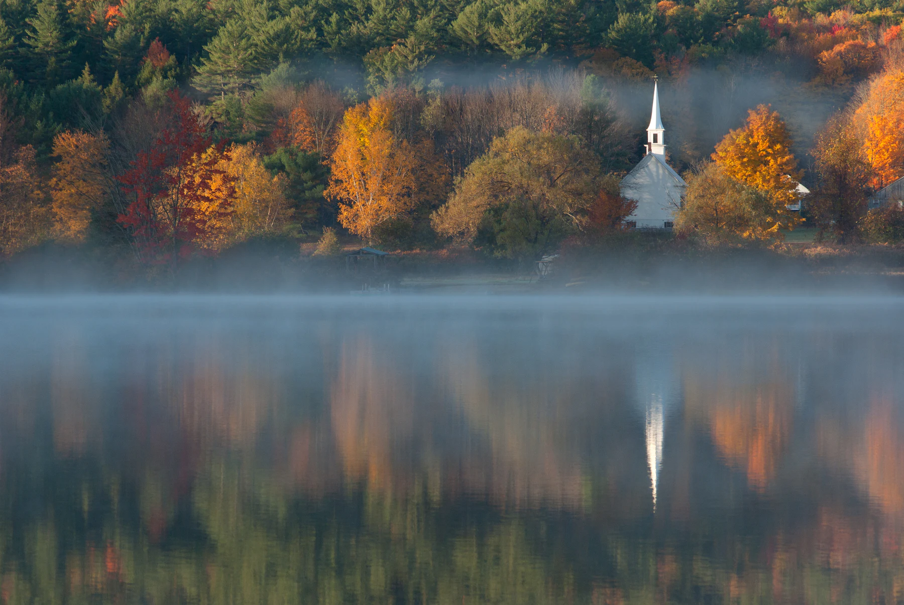 A white chapel among autumn trees, reflected in a misty lake at morning
